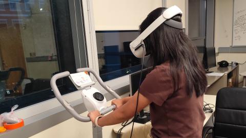 A woman on an exercise machine wearing a VR headset in an office or lab.