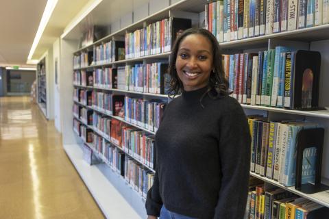 Person standing in a library aisle next to shelves filled with colorful books, wearing a dark sweater and jeans.