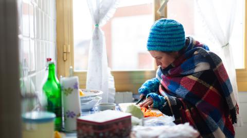 A person wearing a hat and warm clothing prepares food in their kitchen.