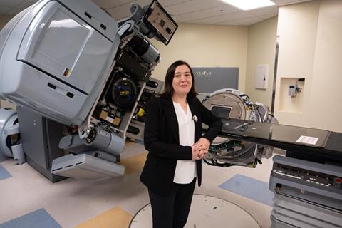 Person standing in a medical treatment room with advanced radiotherapy equipment, including a large Varian machine and treatment table.