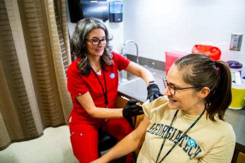 Person in red scrubs administering shot to person wearing tech gold shirt 