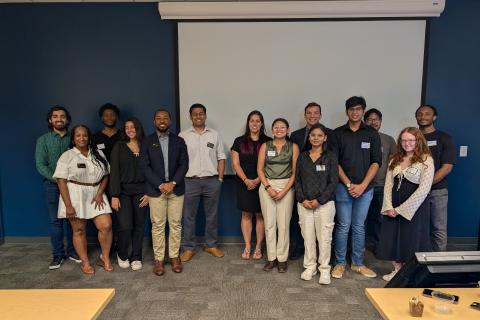 Group of 14 students and recent alumni standing in front of a projector screen in a classroom 