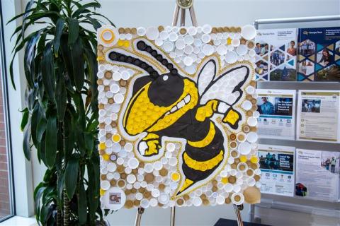 artwork created using plastic lids on display with fliers and plant in background