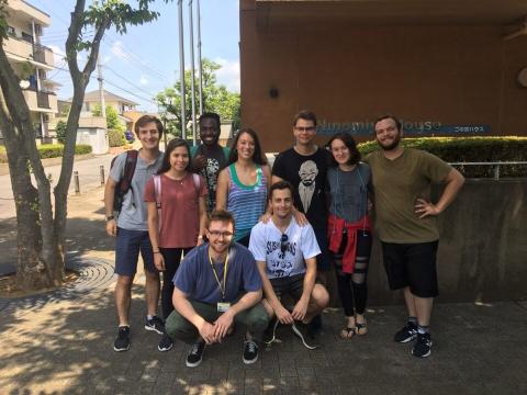 Nine young adults pose together outside a building on a sunny day. Some stand while two kneel in front. They are dressed casually, smiling at the camera, with trees, a sidewalk, and a building entrance sign visible behind them.
