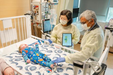Two adults wearing protective gowns and gloves stand beside a hospital crib, using a tablet device while examining an infant lying on the mattress as medical equipment and monitors surround the crib.