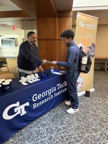 A man behind a table draped with banner reading Georgia Tech Research Institute hands a flyer to a young man.