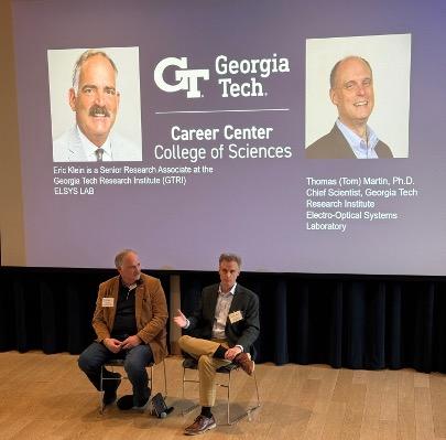 Two men sit in front of a slide featuring their faces and job titles.
