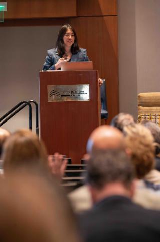 Vanessa Chan speaks at a podium at the Georgia Tech Hotel and Conference Center, addressing an audience. She holds a clicker and stands behind a laptop during a formal presentation.