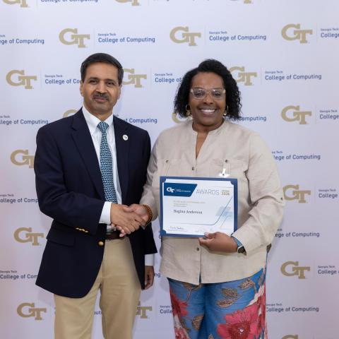 A man and a woman standing next to each other. The woman is holding up a certificate. They are shaking hands.