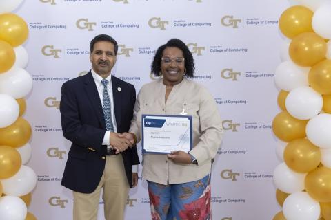 A man and a woman shake hands in front of a step and repeat banner. The woman is holding a certificate.