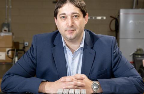 Person seated at a table with hands clasped, wearing a blue blazer and light-colored shirt, with small metal components arranged in front and laboratory equipment visible in the background.