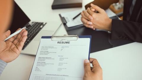 An interviewer holds a clipboard with a resume on it while discussing qualifications with a candidate sitting across the desk.