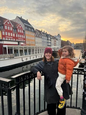 Person stands holding a small child beside a black metal railing along a canal lined with colorful multi‑story buildings. A long white boat labeled ‘LOIVA’ is docked in the water, and additional boats are moored along the right side. The scene is lit by soft sunset light with a cloudy sky overhead and reflections visible on the water.