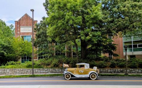 The Ramblin Wreck drives past the Petit Biotechnology Building, a red brick building with large trees in the foreground.