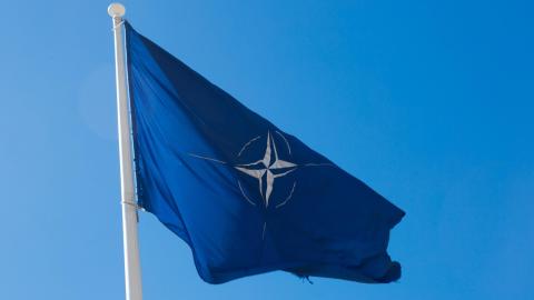 A flag with the NATO logo blows in a clear blue sky.
