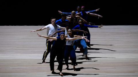 Eight dancers, casually dressed in warm-up clothes, are in a loose group facing the camera their arms outstretched and their legs bent, seeming to stamp their way forward. They are illuminated by a circular spotlight that sets them off from the dark background and pale floor.