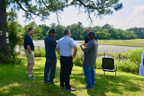 Five men stand in a sunny yard overlooking the marsh in deep conversation. From left to right: South Carolina Aquarium Chief Executive Officer Kevin Mills, John Carr, Chairman of the South Carolina Aquarium Conservation Committee Jonathan Zucker, former South Carolina Aquarium Director of Conservation Albert George, and School of Biological Sciences Professor Joel Kostka.