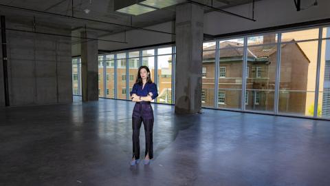Birney standing in an empty building floor looking towards a window. Her hands are crossed in a powerful, standing pose.