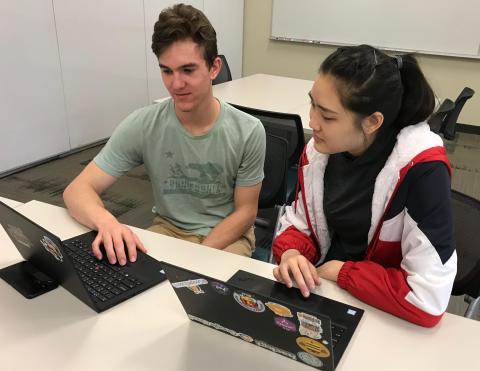 A man and a woman sit next to each other at a table with laptops and talk about their work.