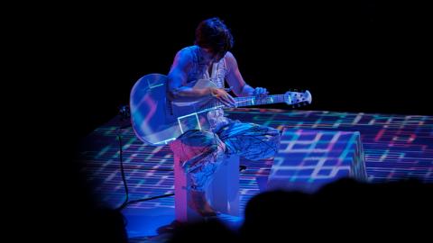 Against a black background, a woman in a silver jumpsuit sits on a chair playing a white guitar. Digital patterns in blue, purple and white are projected on her, the guitar, and the floor around her.