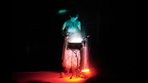 Against a black background, a woman stands at a single drum, Lights shine up on her and the drum, illuminating them in a reds and greens.