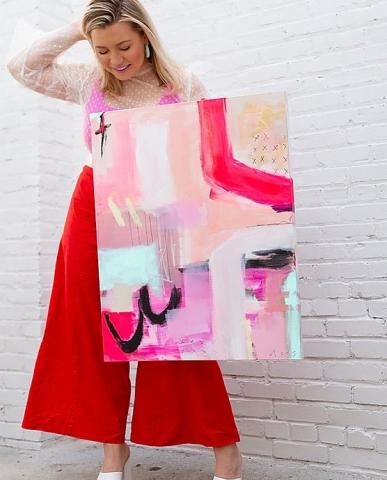 woman wearing a pink top and red billowy pants, standing against a white brick wall.  She holds a large colorful canvas in her left hand, partially covering her body; she holds her hair back with her right hand and looks down.