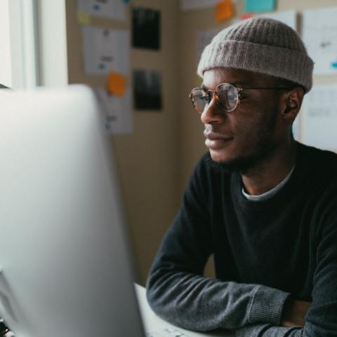 A man in a beanie and long-sleeved t-shirt sits in front of a computer screen.