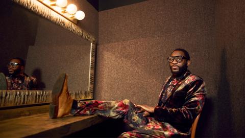 Mwenso wears black frame classes and and a velvet floral suit. He sits in a dressing room in front of a mirror with one leg propped up on the table, while he looks straight at the camera.