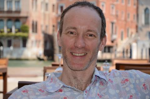 Male figure is seated casually, facing the camera. He is smiling broadly. He is wearing a casual, patterned shirt and is in front of multi-story brick buildings.