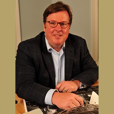 Mr. Russ Todd, from the waist up sitting with his hands folded on a table, smiling, facing the camera