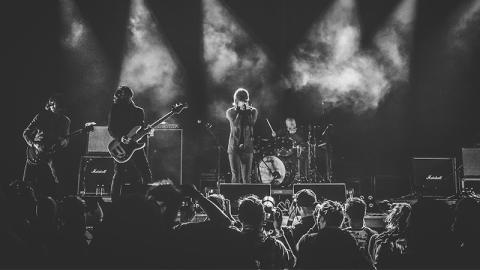 A black and white photograph of musicians on stage, smoke illuminated in the spotlights, the men playing guitar, bass, drums, and singing. Below and in front of them are concert-goers, looking up at the stage.