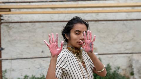 A woman seen from the chest up, standing outside in front of a plain wall.  She holds her hands up, partially covering her face. The palms of her hands are covered in pink paint.