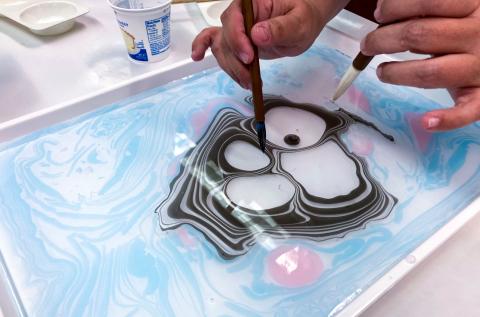 Image of ink floating on water, and two hands holding brushes. The multicolored paint is forming concentric circles like tree rings