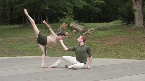 The setting is an outdoors, the woods and grass surrounding a low stage. A male dancer sits, one hand touching the ground, the other gently cupping the female dancer's face. She is posed in a deep arabesque towards the man, her face in his hand and her upper arm and leg extended skyward.