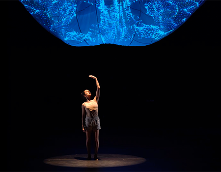 A woman wearing beige shorts and tank top stands alone in the spotlight, her left arm raised above her head as she looks towards her palm. Above her looms an amorphous blue projection. 