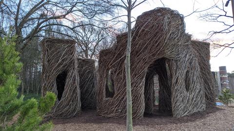 With branches from green trees in the foreground and a tall building in the background there are structures made from woven saplings, with arched openings to walk through