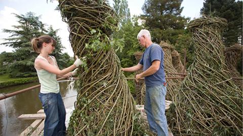 A woman and a man are outside, dressed in warm-weather work clothes. They are working with branches and saplings, winding them around a 10-foot high sculptural installation made entirely of natural materials. Similar structures are visible in the background.