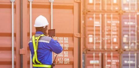 Port docker wearing hard hat and safety vest, standing in front of freight containers looking at supply chain trends on tablet