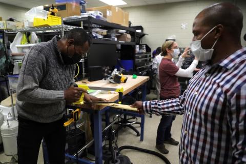 Two men saw off a piece of wood while a woman stands in the back talking to another person.