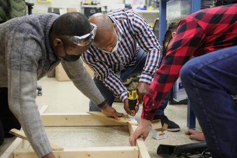Three men are holding down a 2 by 4 wood frame and using a screwdriver to nail down a part of the frame to secure it.
