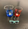 Two large beakers — one filled with blue water and one filled with red water — behind a small beaker filled with purple water. Small safety glasses are in front of the beakers.
