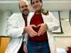 Christina Ragan, currently pregnant, and Zachary Grieb pose together in lab coats, forming a heart with their two hands over Ragan's abdomen.