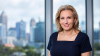 Laura Taylor, a blonde woman with a fair complexion, stands in a navy dress in front of a large window with a blurry skyline of Atlanta visible through the window.