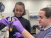 Two people wearing gloves and eye goggles look at substances in a test tube.