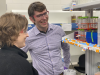 Two people wearing protective eye goggles chat in a lab, looking at test tubes.