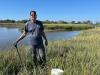 A man in a blue shirt holds a shovel in a salt marsh. 