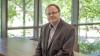 Professor Michael Hoffmann poses for the camera in front of a window wall showing verdant summer foliage.
