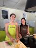 Two young women wearing aprons pose in front of a cooktop