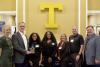 Seven people pose with an award in front of a giant letter T.