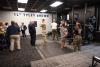 A group of people, some in military uniforms, talk inside an exercise room with dark walls, carpet, and ceiling tiles and a sign on the wall reading "1LT Tyler Brown Fitness Facility."